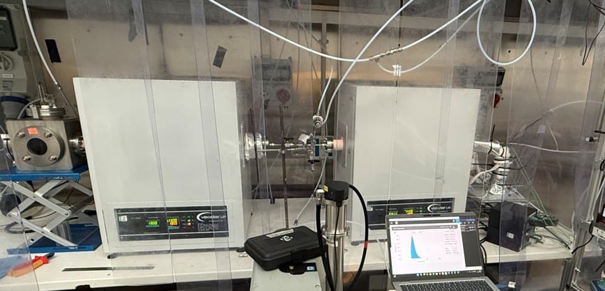 Photograph of the physical experimental rig: twin tube furnaces, gas delivery lines, and a laptop showing live particle-size data.