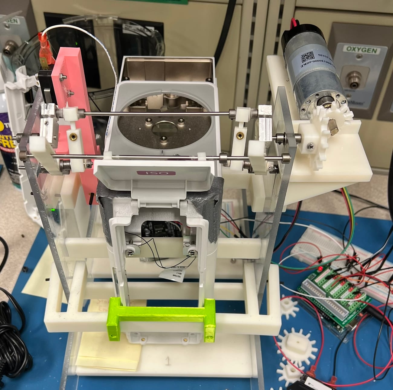 A benchtop cam-and-spring fixture: a DC motor drives a cam that cycles a vaporizer cover-door mechanism, with Arduino control.